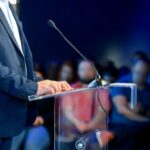 A debater stands at a microphone stand, hands resting on the podium while reviewing notes. In the background, an out-of-focus crowd sits attentively