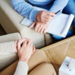 a dcotor and patient sit across from each other in arm chairs, only their hands can be seen. The doctor has a notepad and pen in their hand. The patient's hands are clasped together.