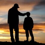 A father and son stand side by side on the shoreline, silhouetted against the horizon as they share a moment together
