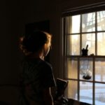 In a dimly lit room with no lights, a girl gazes out of a window. The soft glow from outside gently illuminates her hairline