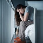 woman sitting on the side of a bed looking sad, holding her head in her hands