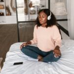 A woman sits on a bed with crossed legs, wearing headphones and immersed in meditation