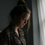 A young woman in a dimly lit room, near a window, her gaze directed downwards, conveying a sense of sadness and contemplation.