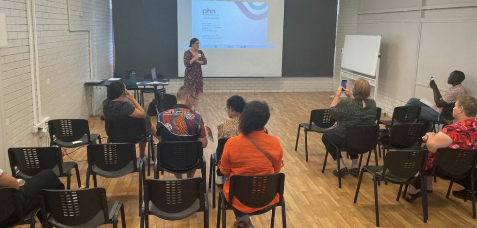 A group of mental health professionals are sitting on chairs and watching a presenter speak at the front of the room in front of a powerpoint presentation