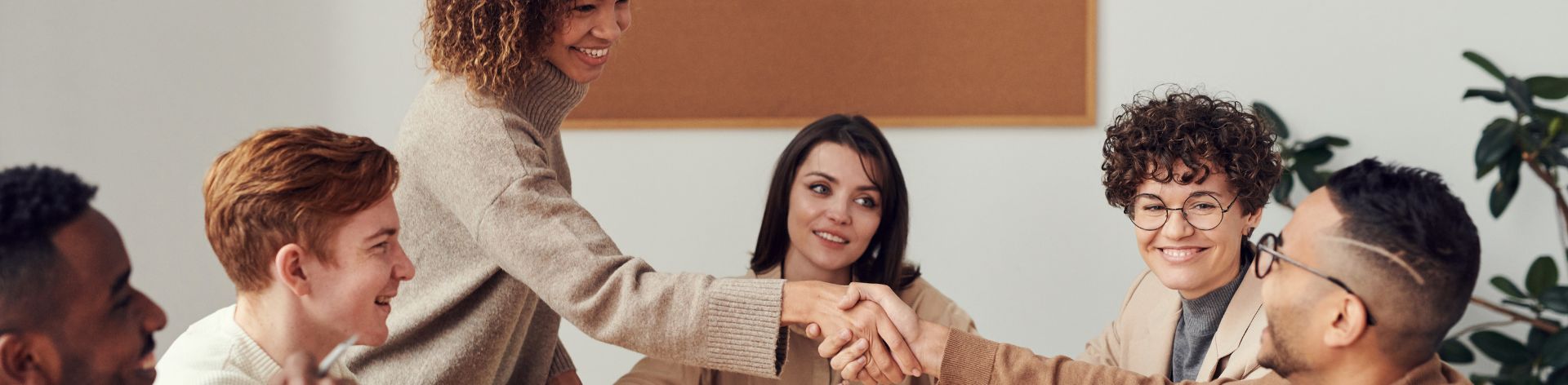 A group of people sit around a table in an office. One woman is standing and reaching across the table to shake hands with a man.