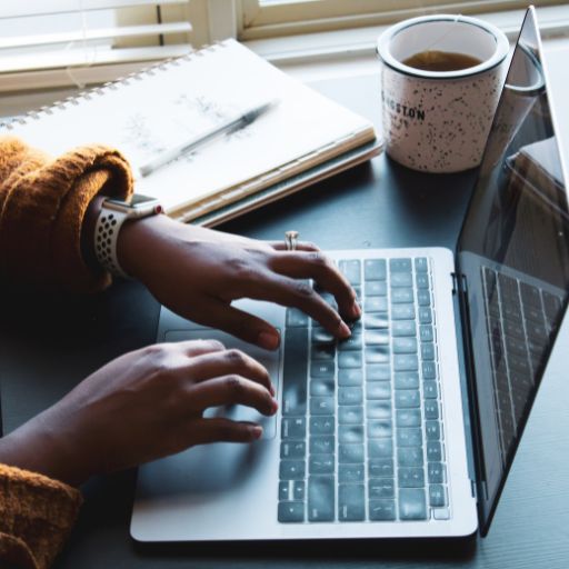A laptop sits on a desk with hands typing on it