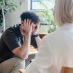 man at a psychologist appointment holding his head in his hands. His psychologist sits in front of him on a chair