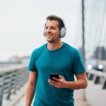 A man is walking outdoors along a bridge, wearing headphones and holding his phone