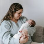 A mother sits on the couch while holding and looking down at her baby.