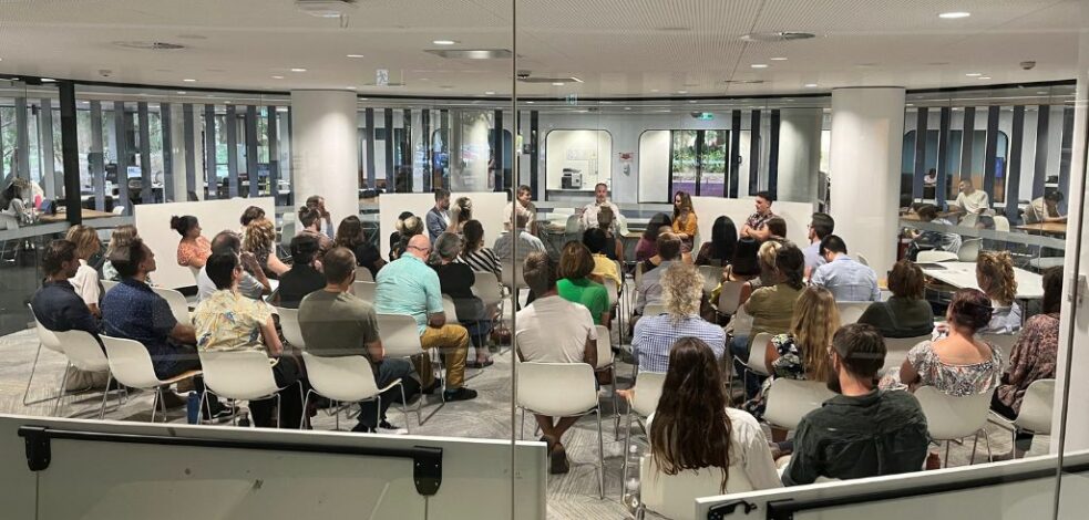 A large group of mental health professionals sit in chairs gather around to focus on a presenter at the front of the group.