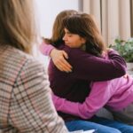 Two women share a loving embrace while sitting in a room with a therapist.