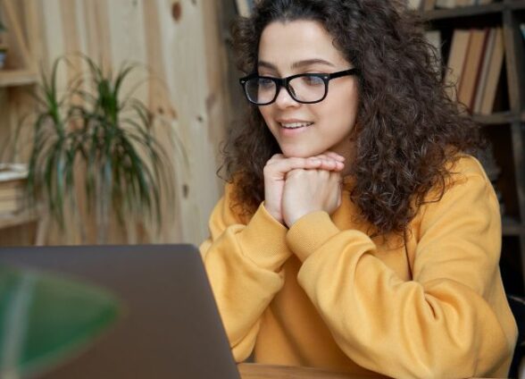A woman sits at a desk with hands under her chin, paying attention to a laptop screen and smiling