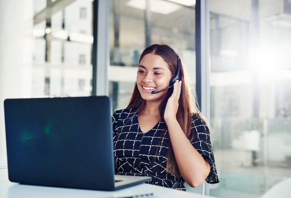A woman sits at a desk working on a laptop, speaking into a headset and smiling