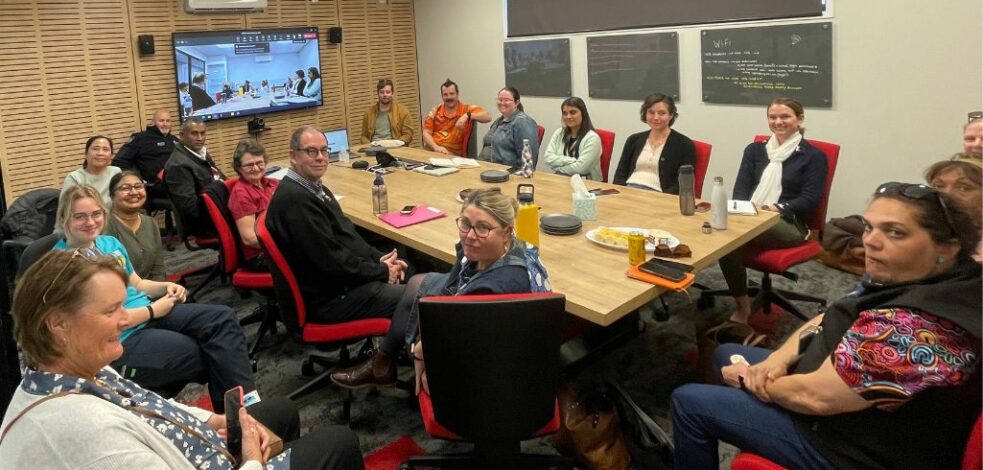 A group of mental health professionals sit around a table in a board room with online attendees on a TV screen, smiling at the camera for a photo.
