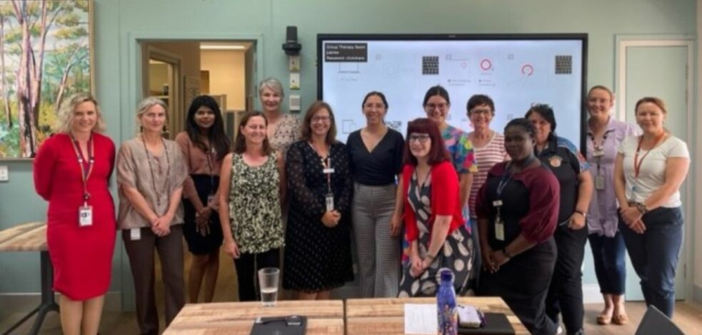 A dozen practitioners stand at the front of a room smiling for the photo, in front of where a presentation was held.