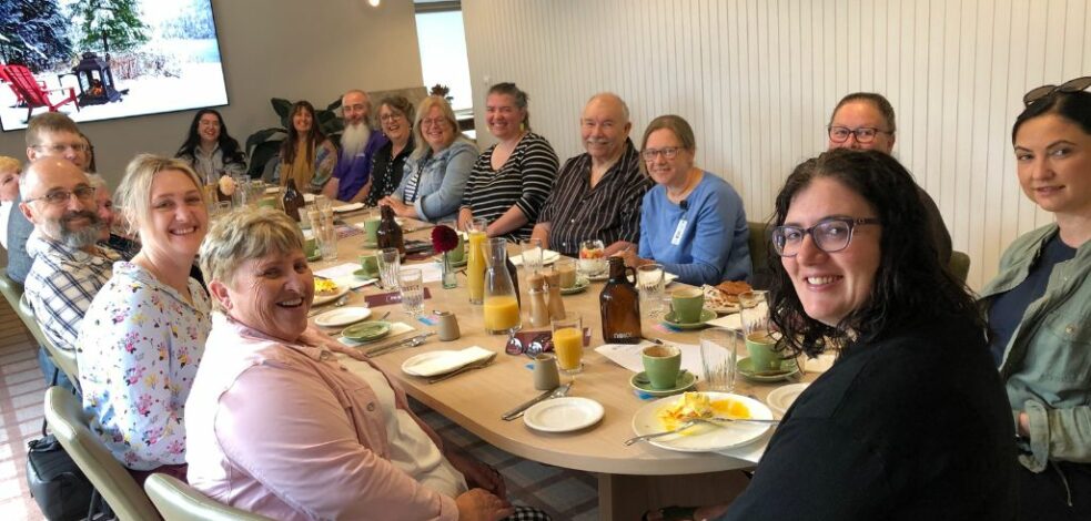 A group of 15 practitioners sit around a large cafe table eating breakfast and smiling.