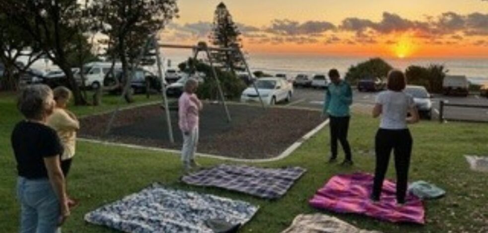 A group of five practitioners stand in a park at sunrise, each with a picnic rug to practice yoga and movement while networking.