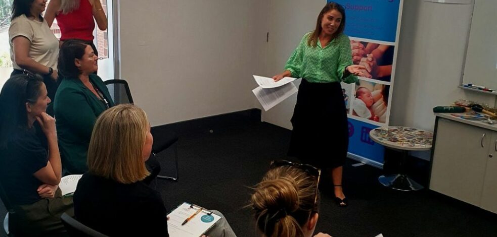 A woman stand at the front of a group of practitioners giving a presentation about perinatal mental health and support.
