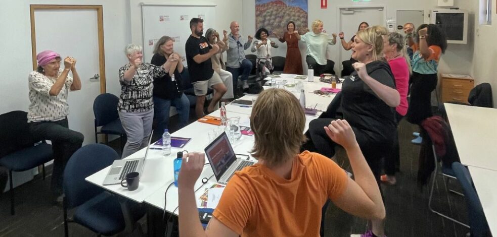 A group of practitioners in a meeting room stand up around a table and participate in a physical exercise.