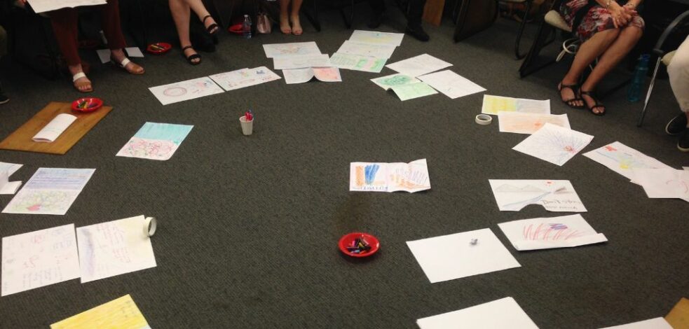 The feet of a group of practitioners can be seen sitting in a circle with many pieces of paper and drawings scattered on the floor in the middle for viewing.