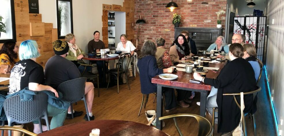 A group of practitioners sit on various table in a cafe talking and networking.