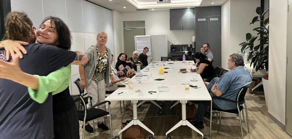 A group of practitioners sit around a meeting room table preparing to begin the meeting. Two people are hugging in greeting.