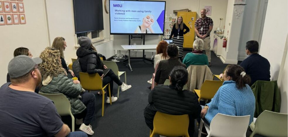 A group of a dozen practitioners gather facing two presenters at the front of a room next to a presentation slide titled 'Working with men using family violence'.