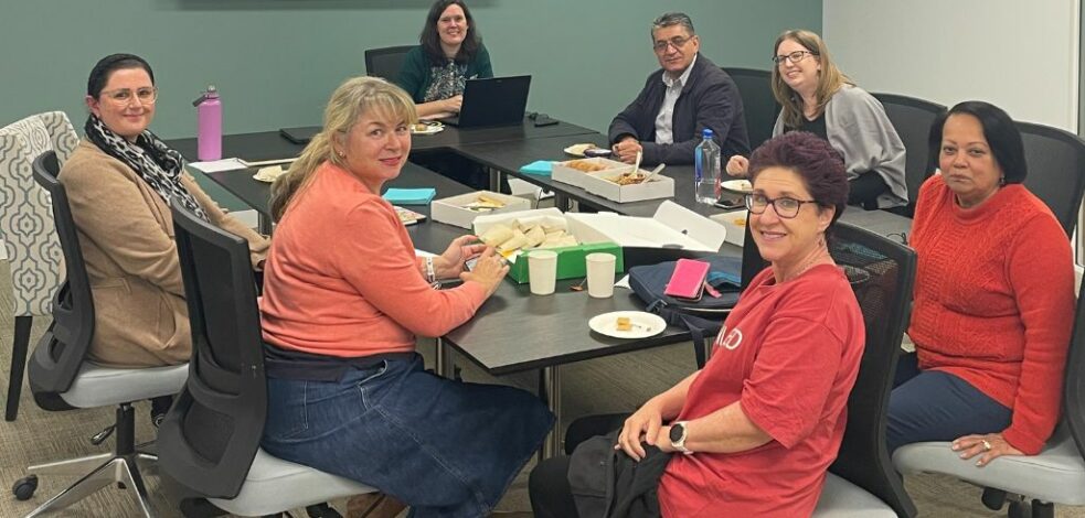 A group of seven practitioners sit around an office meeting room table, sharing food and smiling for the camera.