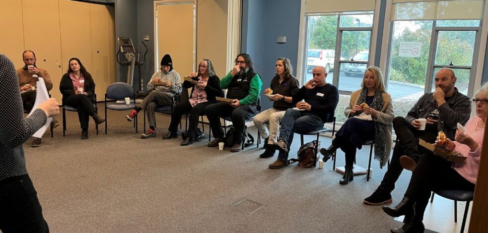 A group of practitioners sit on chairs arranged in a semi-circle, eating breakfast while listening to a presenter speak.