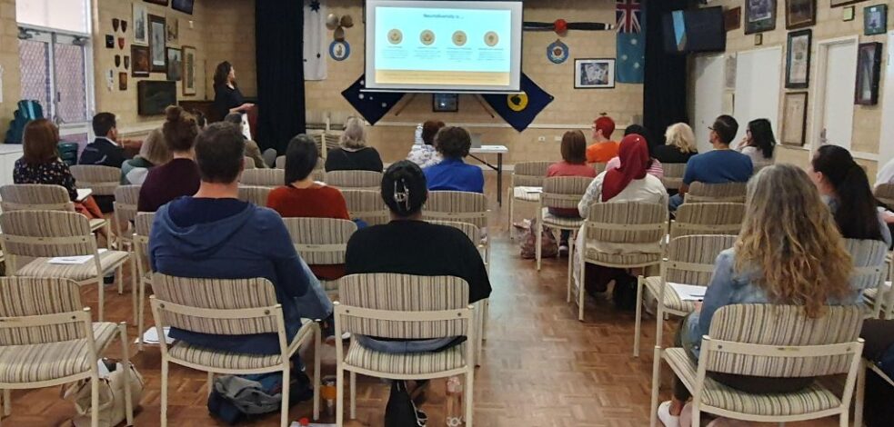 A group of people sit in a room facing a presentation at the front.